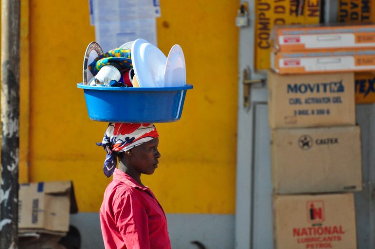 A woman walks with a bucket of dishes atop her head