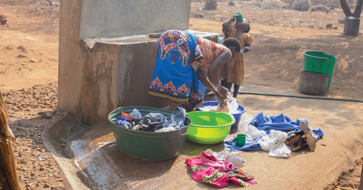 A woman cleans her clothing in water bins while a child drinks safe water from a cup