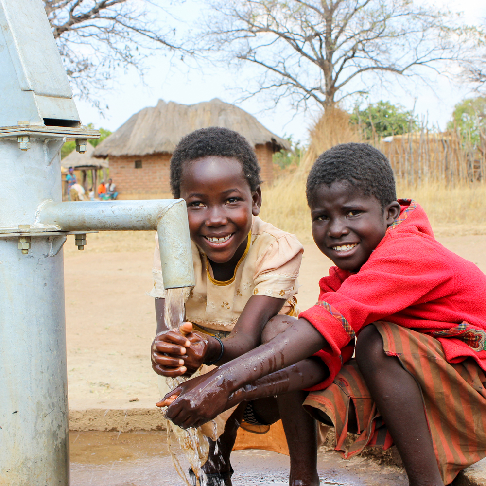 2 boys smiling at the camera while they wash their hands using the new water pump installed in their community