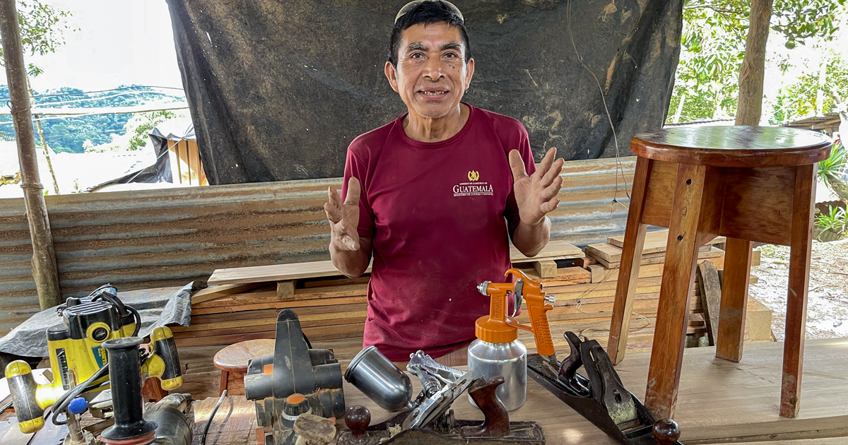 Don Fidel, a Guatemalan man, speaking to the camera while working in carpentry. He is surrounded by tools and appears to be working on a piece of furniture.
