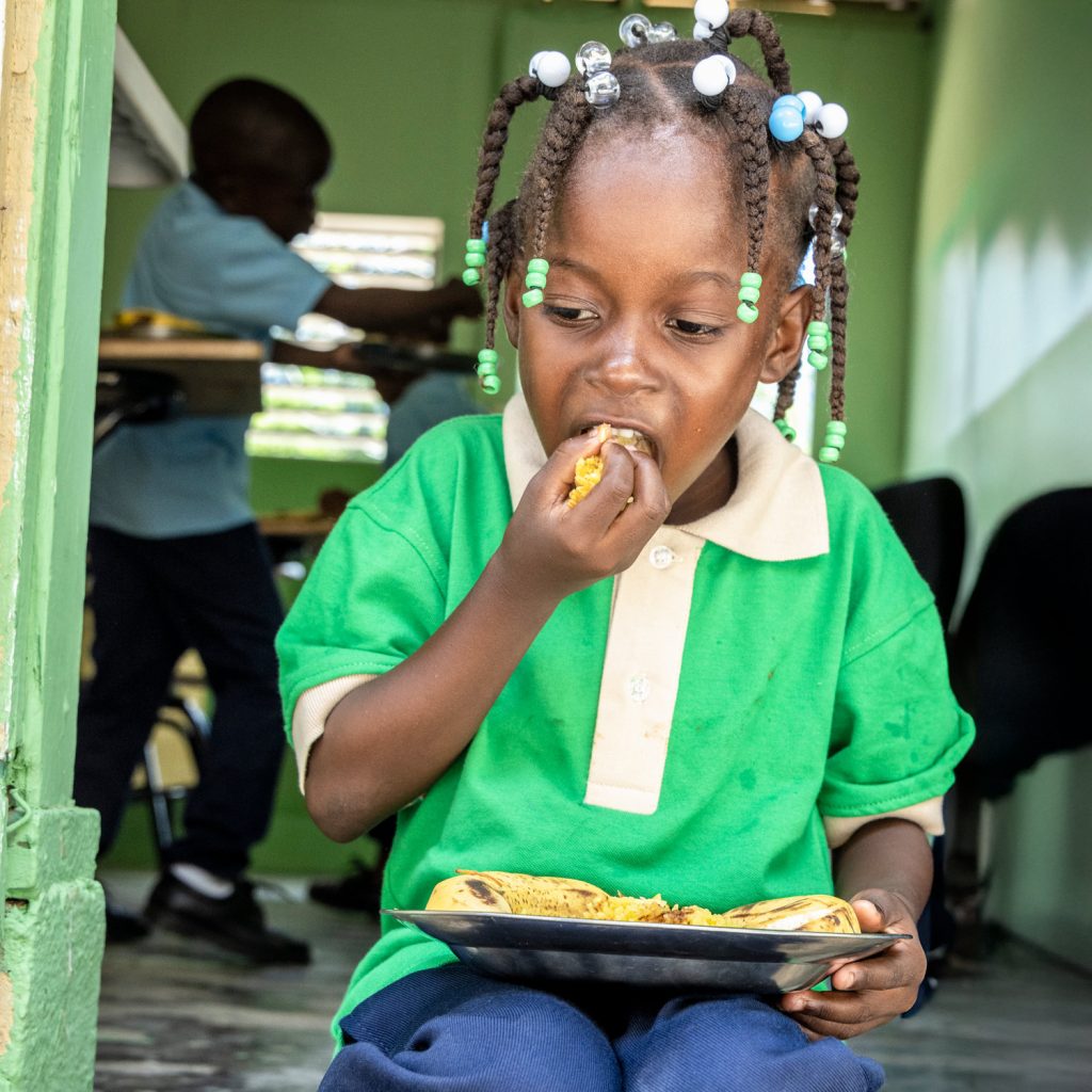 Girl who lives with her family in the Milagrosa batey, Dominican Republic, sitting on a doorstep in her uniform, with a plate of food on her lap, eating.
