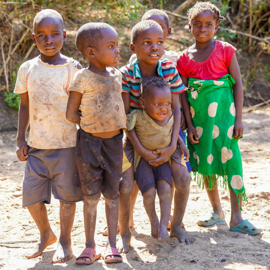Group of children in a remote Zambian village standing on dirt ground with vegetation behind them, some looking directly at the camera.