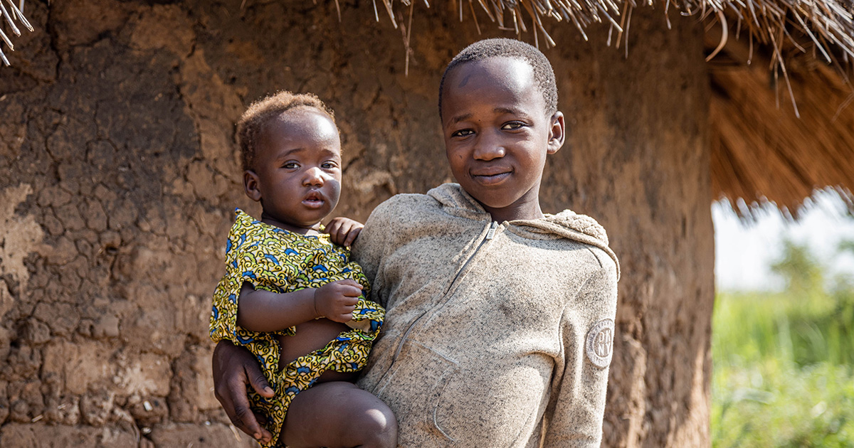A young girl holds a toddler on her hip in front of their Gulu home.