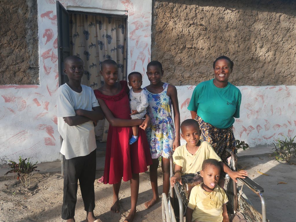 A Kenyan family standing together in front of a house. In front of the group, there are two boys: a younger one in a worn-out wheelchair, with his older brother standing behind it, supporting his arms on the arms of the chair. They are all smiling, posing for the photo.