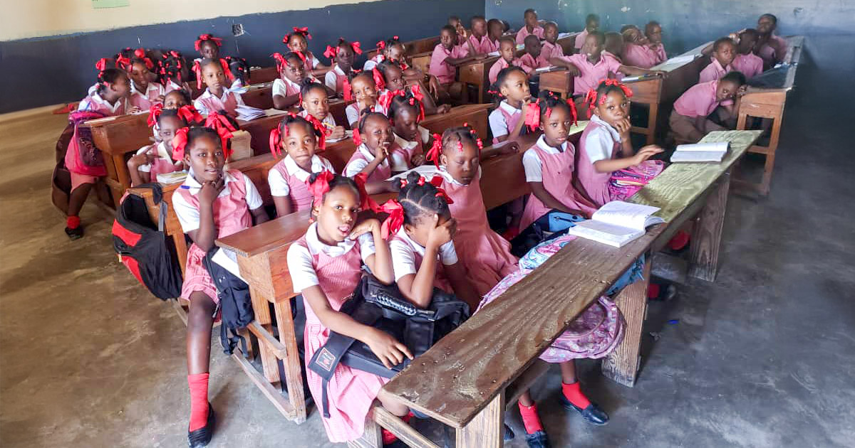 A classroom in a Haitian school with children in uniforms sitting closely together on shared wooden benches, some looking at the camera while others interact with each other.