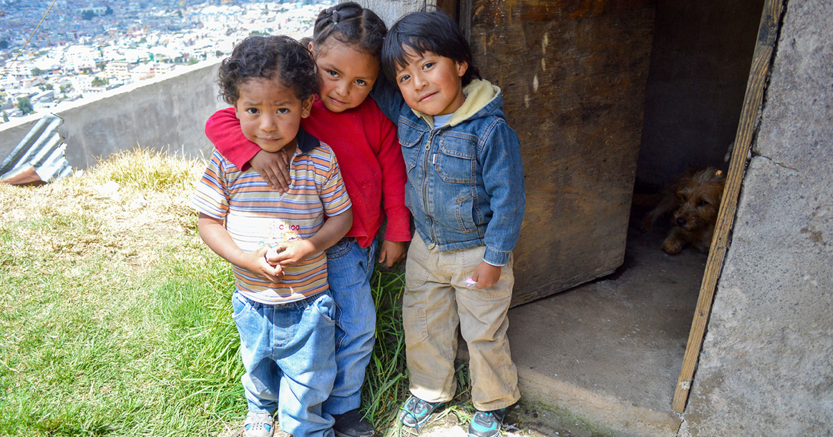 Three Ecuadorian children outside their modest home with a view of the town, suggesting it's on a hill.
