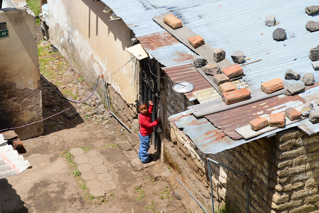 Looking down on a hillside slum in Ecuador, a young girl stands at the doorway of a tin-roofed home.