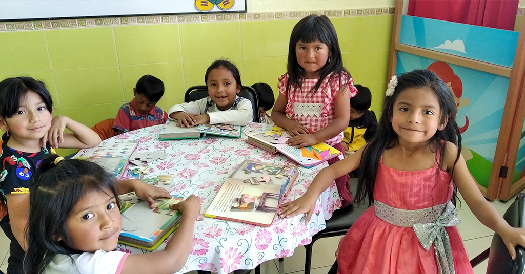 A group of children in Ecuador engage with books, as five girls at the front table pause to look at the camera.