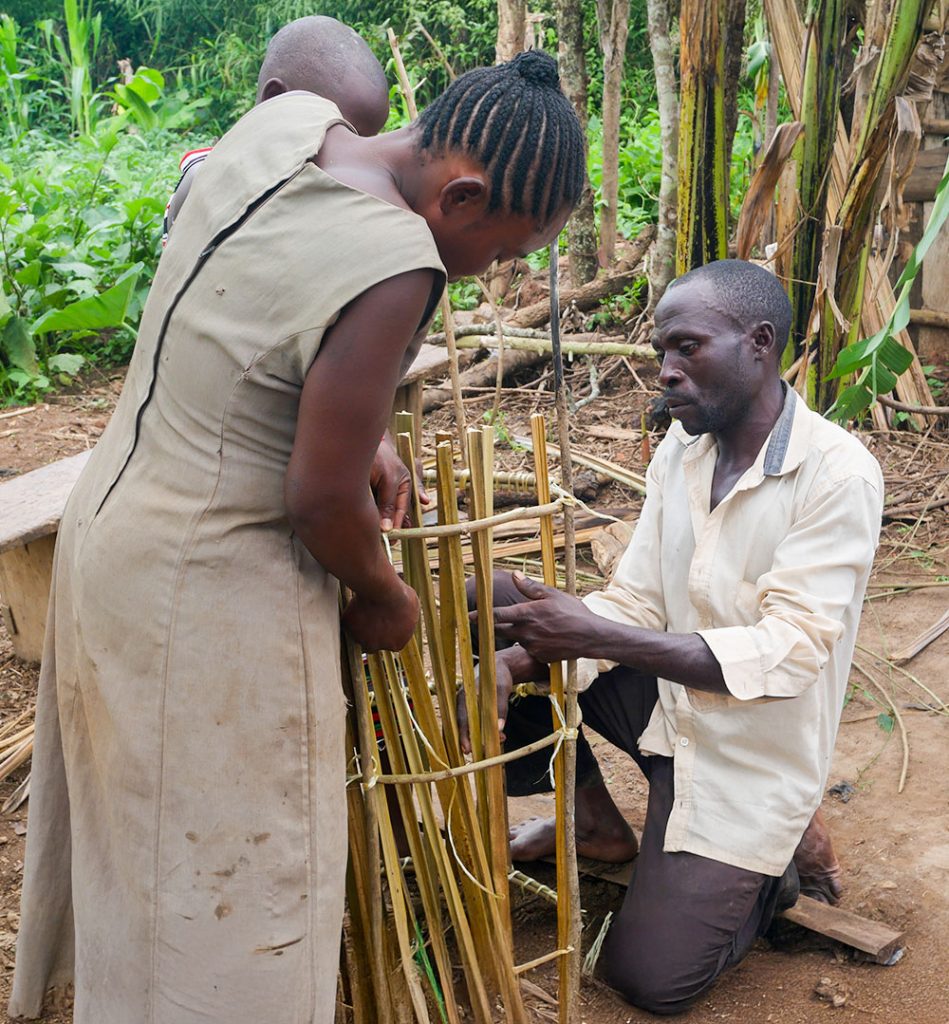 A man kneels beside a standing woman as they weave bamboo poles into the frame of a traditional beehive.