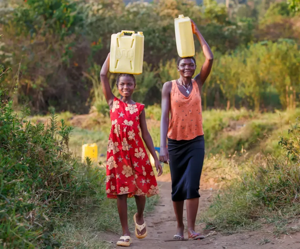 Sabella, a young Ugandan girl in a red dress, and another woman carry water jugs on their heads through a rural, dusty landscape.