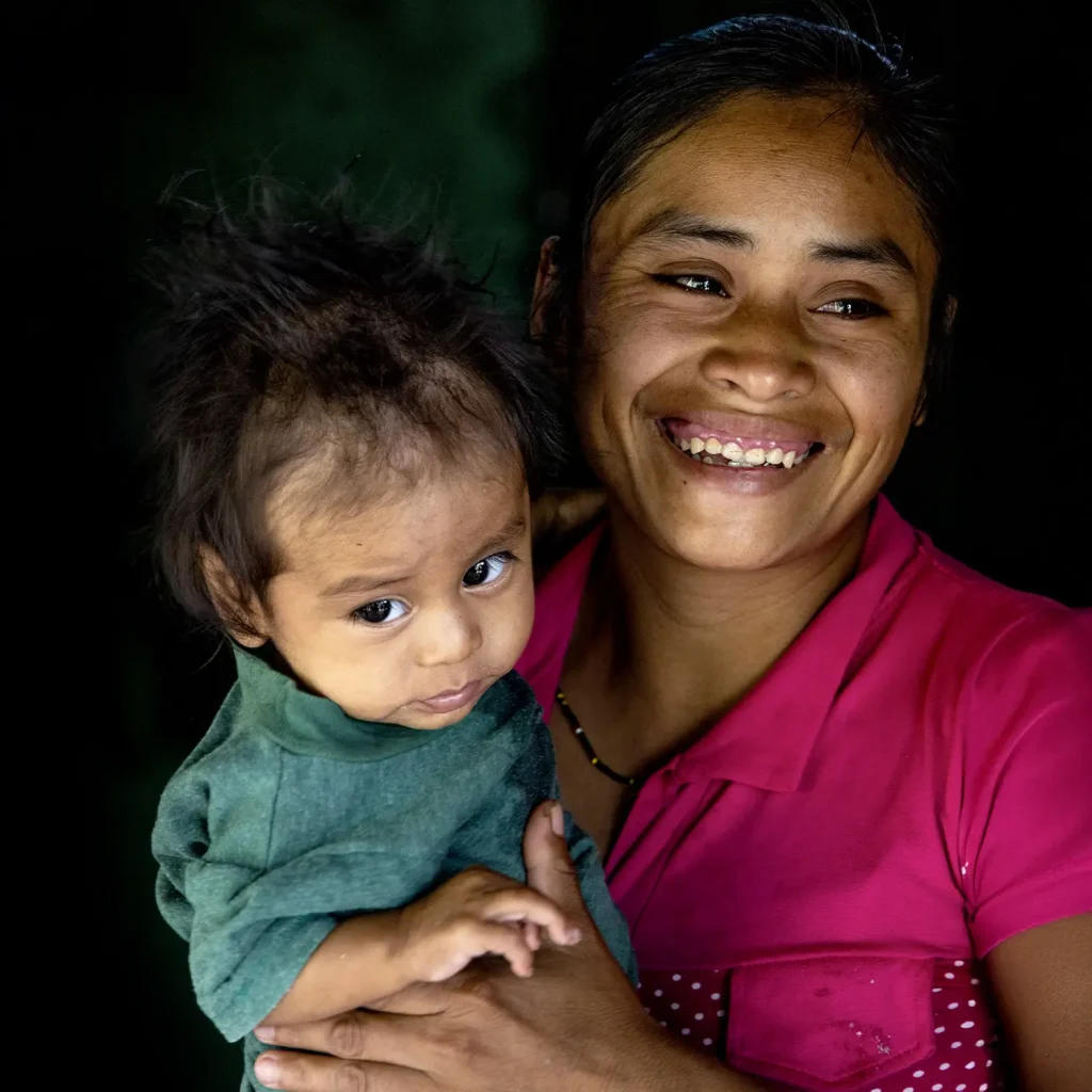 Guatemalan mother in pink holds her baby in green, smiling joyfully while the child looks at the camera.