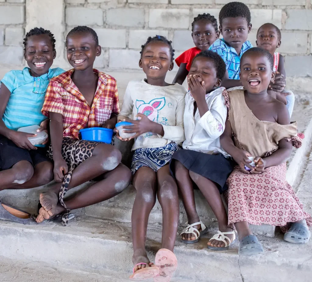 A group of children sit together on a step inside a Fellowship Hall in rural Malawi. They smile and laugh as they hold bowls of food, their joy and friendship filling the scene.