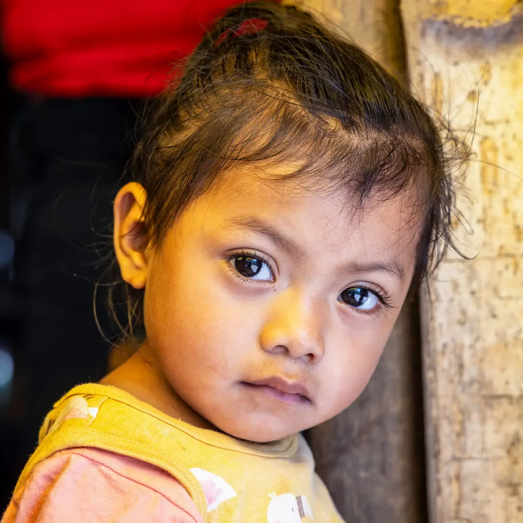 Young girl in La Jigua, Zacapa, Guatemala, gazes softly at the camera while standing near a wooden wall. She wears a yellow tank top over a pink shirt.