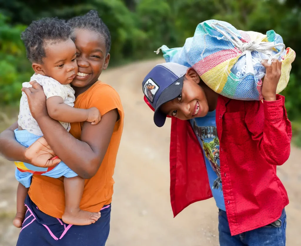 Smiling girl holds a baby while standing beside a boy carrying a colorful sack over his shoulder on a dirt road surrounded by greenery.