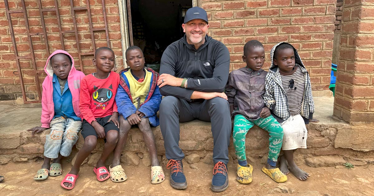 Zach Oles sits with a group of children from a remote African village, smiling together outside a brick building.