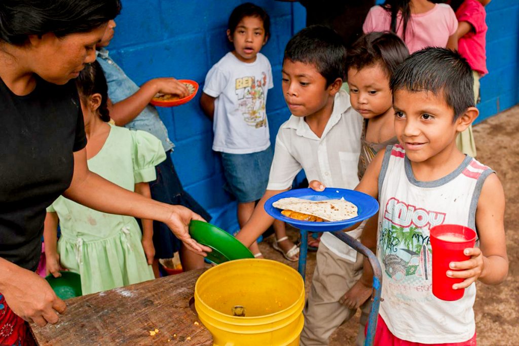 Smiling young boy holds a plate of food and a red cup while waiting in line with other children as a woman serves meals