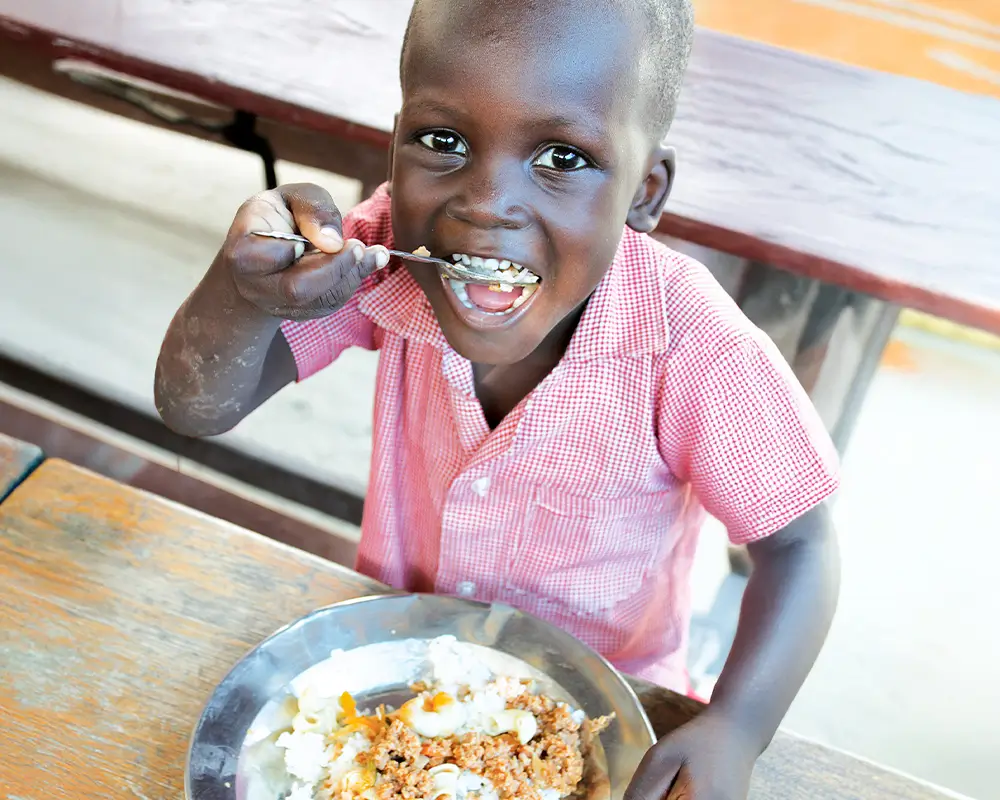 Young child in a red-checked shirt eats from a bowl at a wooden table, smiling at the camera.
