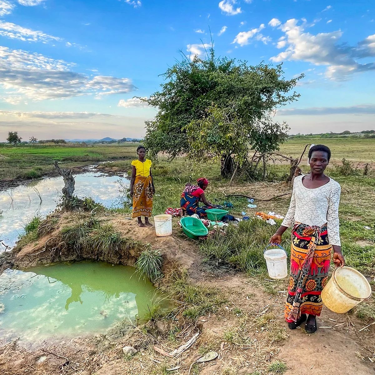 In a remote Zambian village, a woman carries buckets away from a small, polluted pond where others gather to wash.