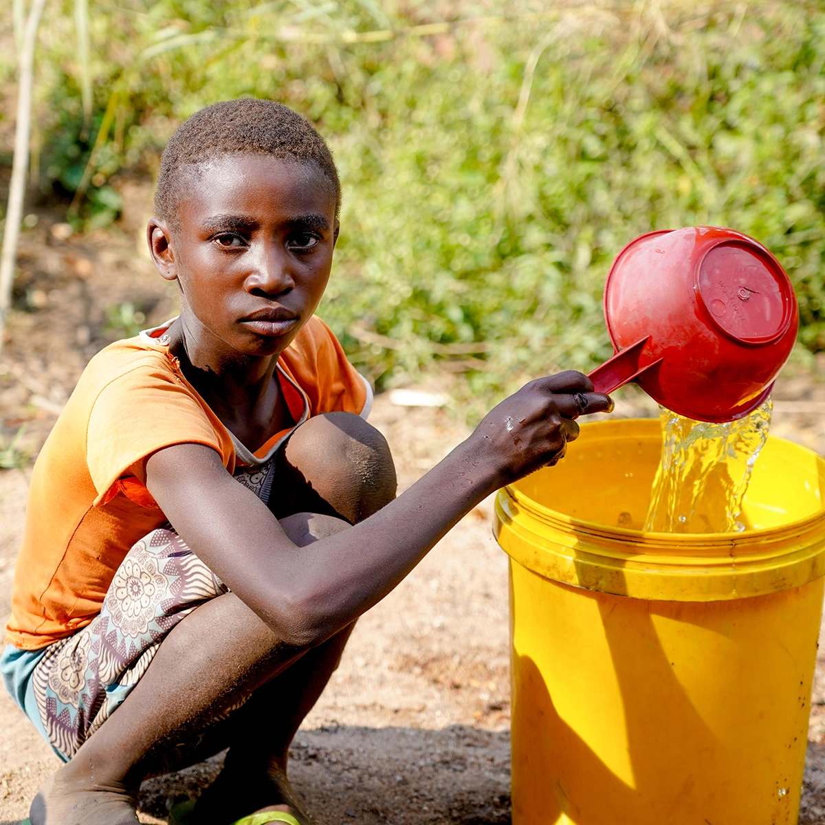 Young boy kneels beside a yellow container, pouring water from a small red cup outdoors.