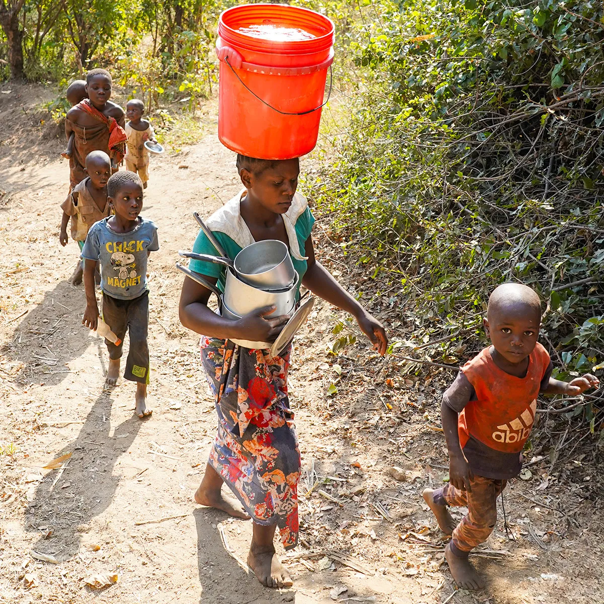 Woman carries a red bucket of water on her head while children walk beside her along a narrow dirt path.