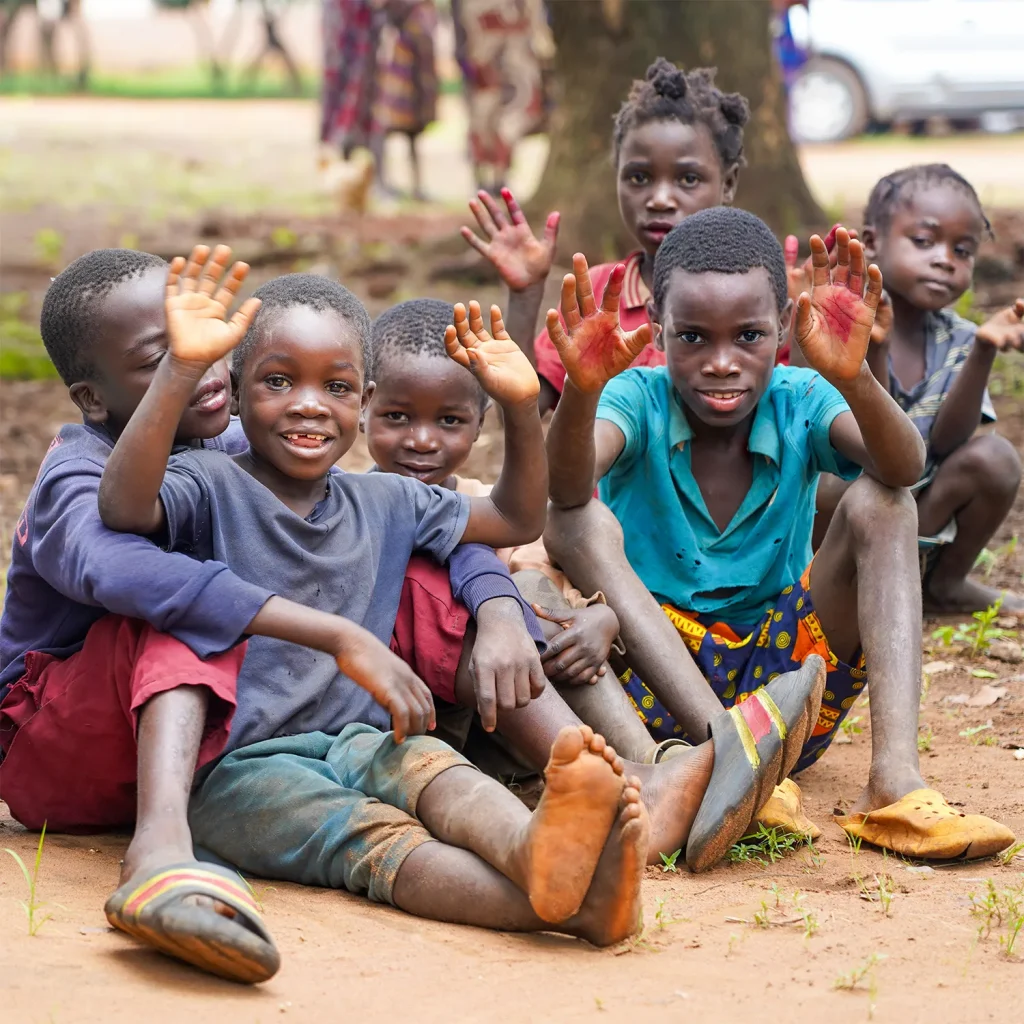 Group of children sitting outdoors on the ground smile and wave at the camera with raised hands.