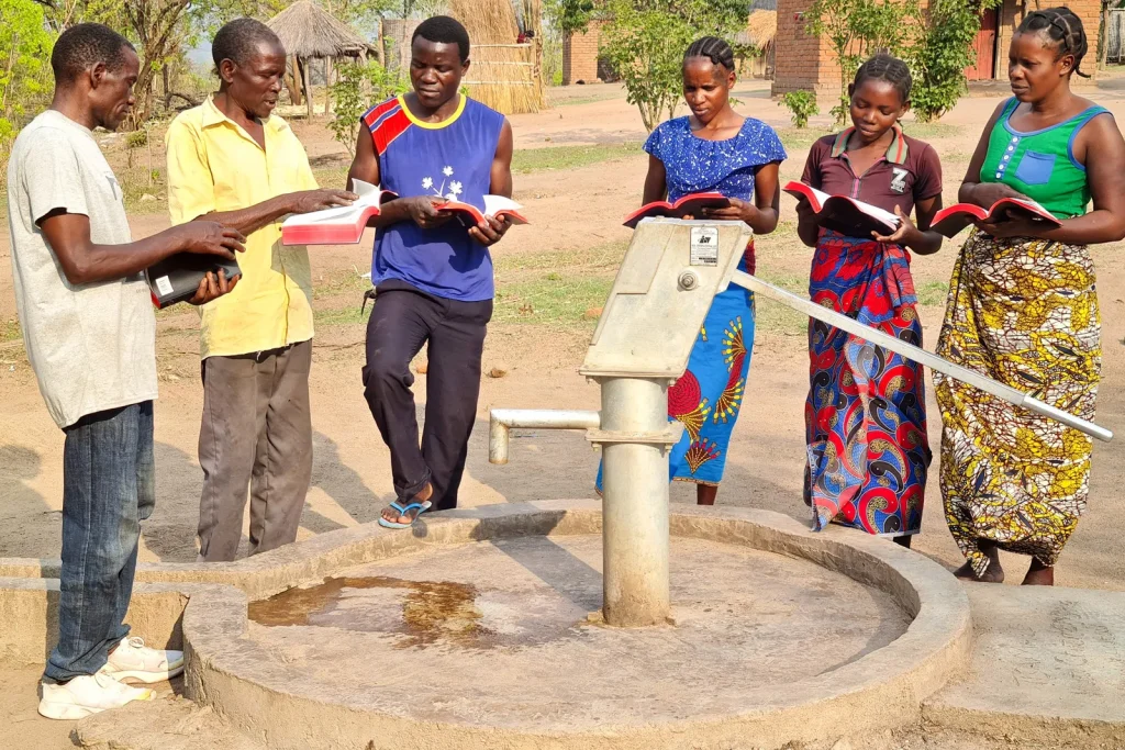 Group of adults in a rural community standing around a hand pump well, holding Bibles.