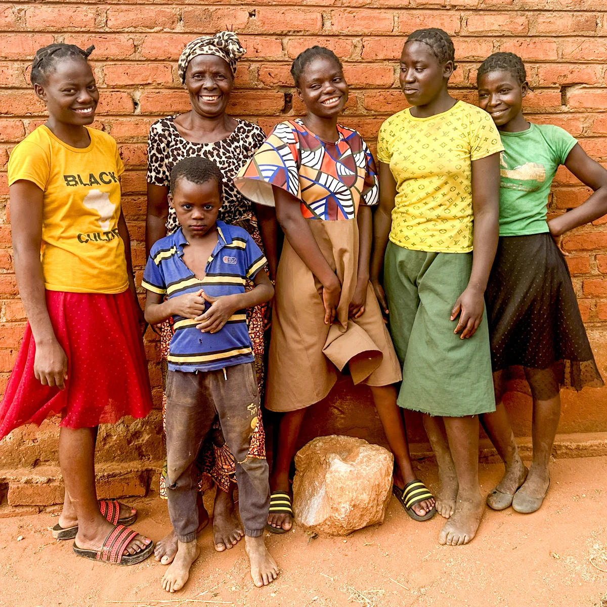 A smiling woman stands with four teenage girls and a young boy against a brick wall, all looking at the camera.
