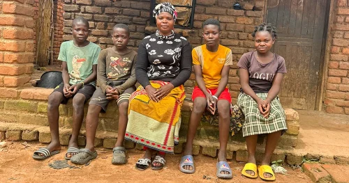 Woman and four children sitting together on a bench outside a brick home in a rural village.