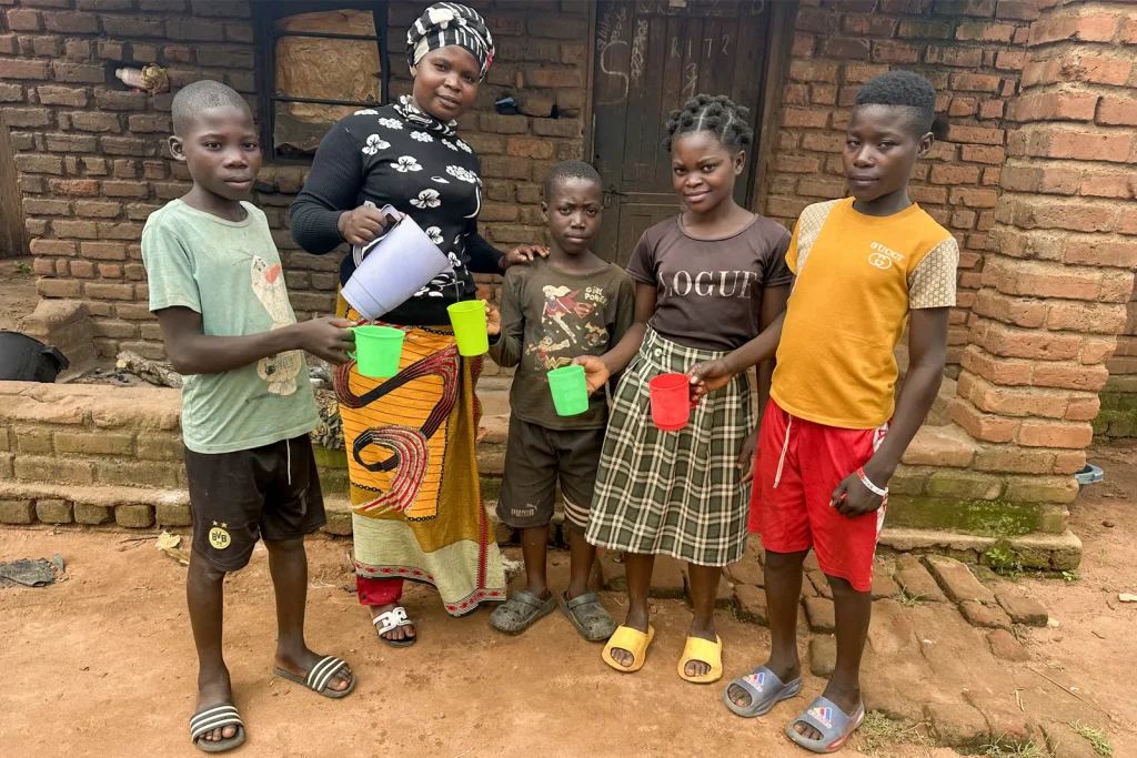 Woman pours drinking water into cups held by four children outside a brick home.