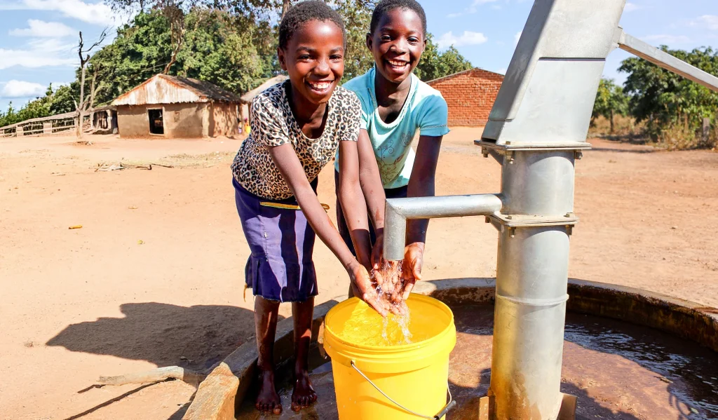 Two smiling girls fill a yellow bucket with water from a hand pump in a sunlit rural village, with small houses and trees in the background.