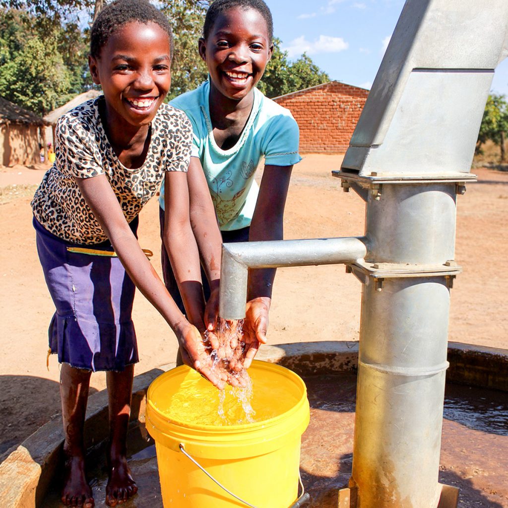 Two smiling girls fill a yellow bucket with water from a hand pump in a sunlit rural village, with small houses and trees in the background.