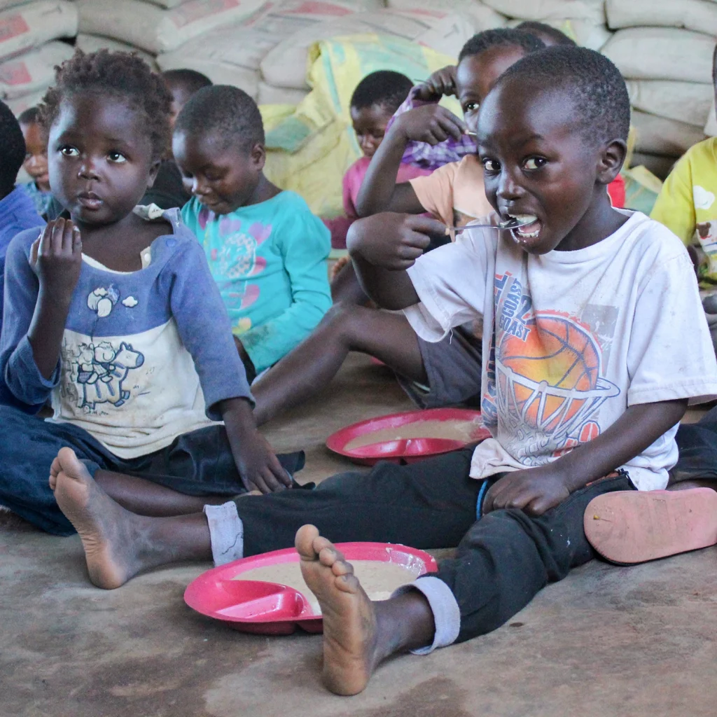 Children in Malawi sit together eating a simple meal, with a smiling boy in the foreground holding a spoon.