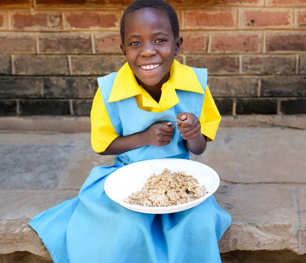 9-year-old girl in a school uniform sitting outside a brick building, smiling and holding a plate of food.