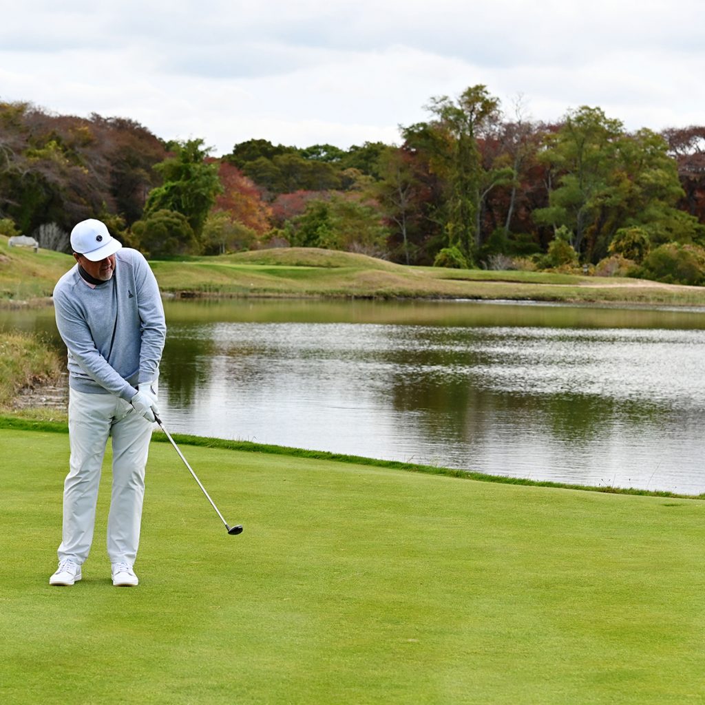 Golfer standing on a green near a water hazard, holding a club and looking toward the course.