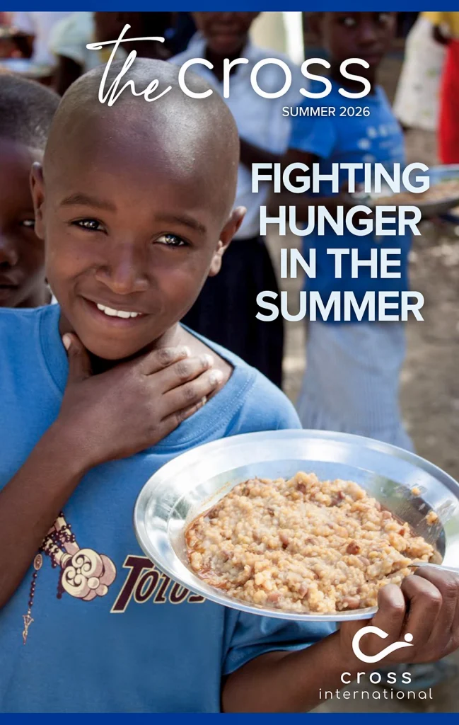 Child smiling and holding a plate of food, with other children in the background, featured on the cover of The Cross Magazine, Summer 2026 issue.