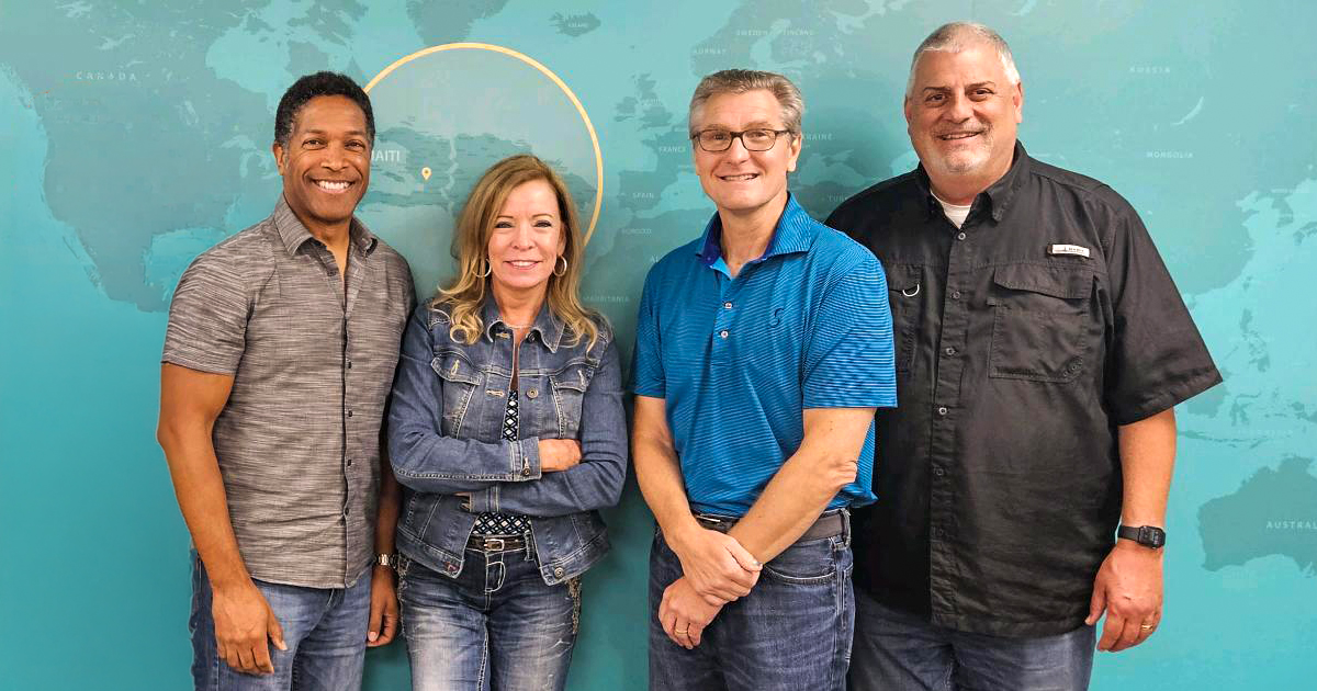 Four adults standing side by side in front of a world map background, smiling at the camera.