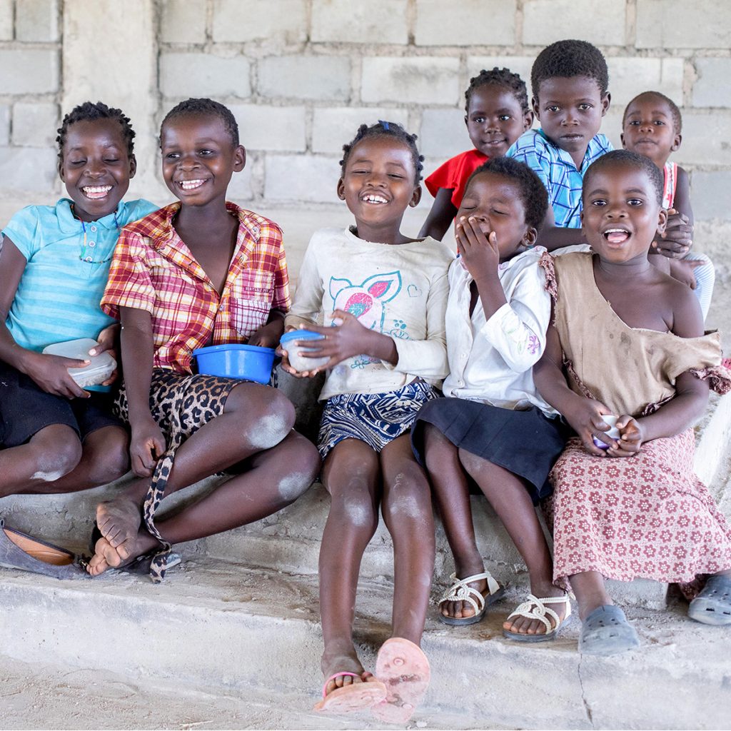 A group of children sit together on a step inside a Fellowship Hall in rural Malawi. They smile and laugh as they hold bowls of food, their joy and friendship filling the scene.