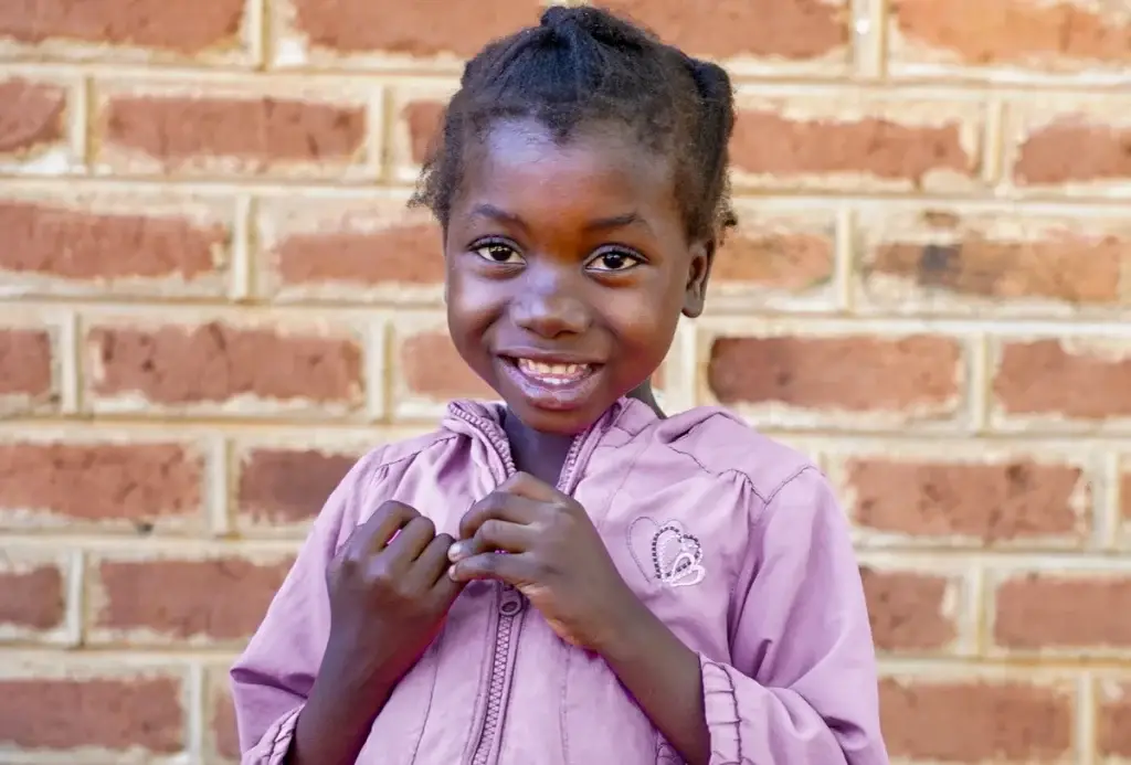 Young girl smiling at the camera in front of a brick wall, holding her fingers near her chest.
