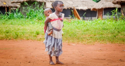 Young girl standing barefoot on a dirt path, carrying a baby on her back near thatched-roof homes.