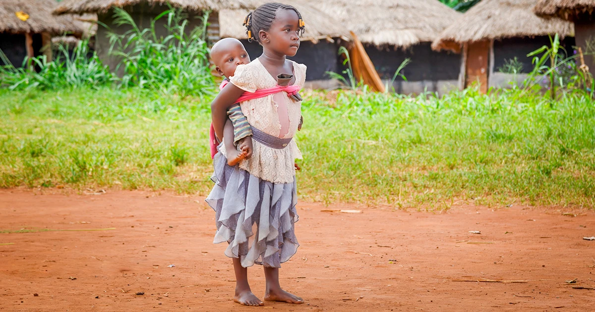 Young girl standing barefoot on a dirt path, carrying a baby on her back near thatched-roof homes.