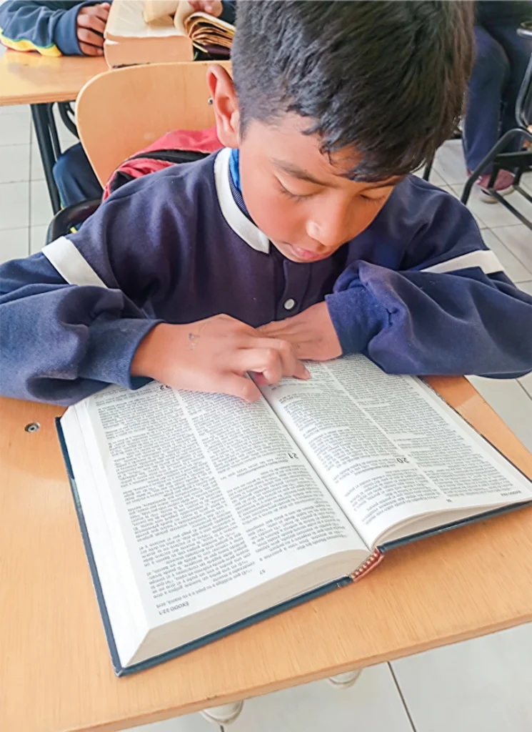 A schoolboy reads the Bible at his desk in a classroom.