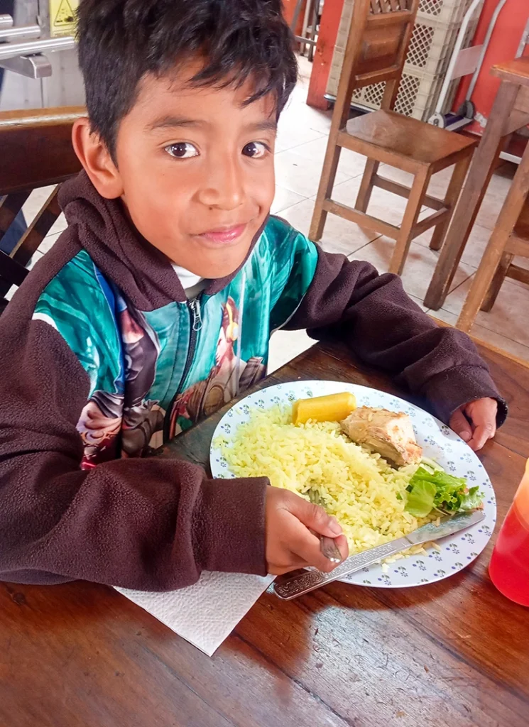 Boy sits at a table holding a plate of food while looking at the camera.