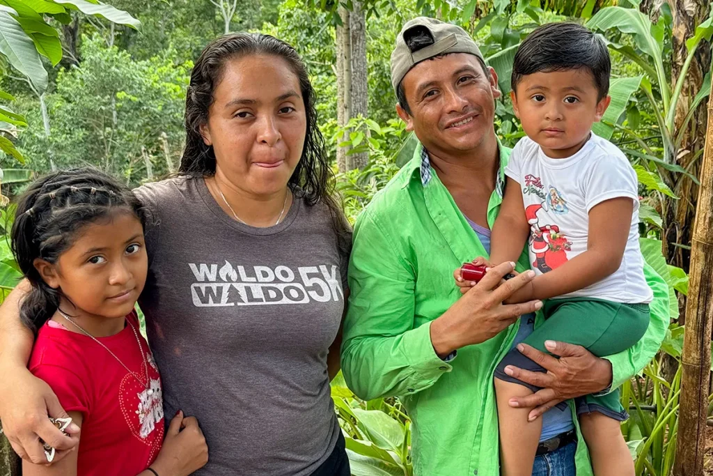 A family of four stands together outdoors, surrounded by vegetation.