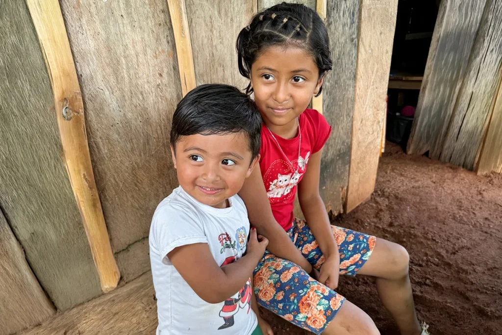 A young boy and girl sit together outside a wooden home.