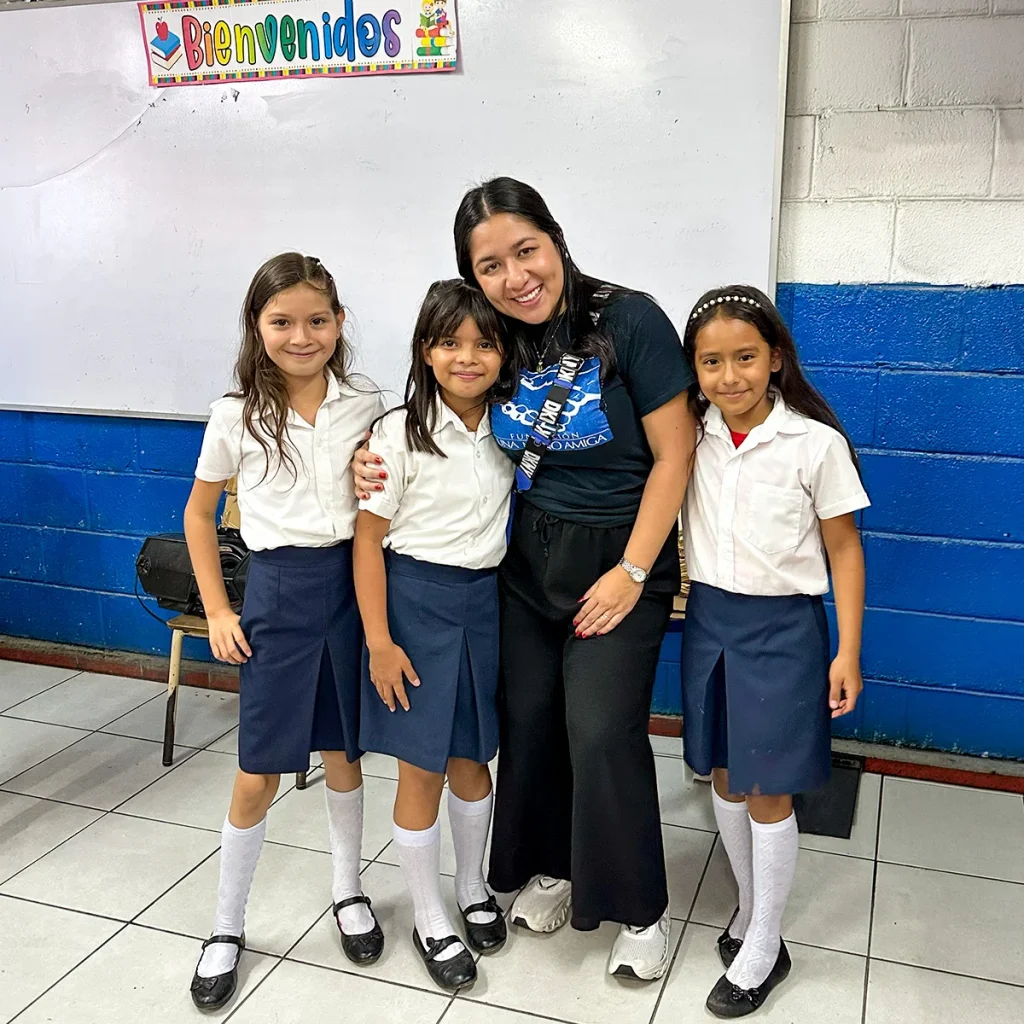 Three schoolgirls in uniform stand with a woman in a classroom.