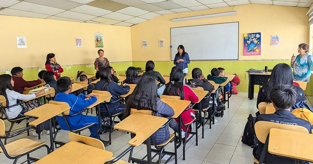 Students seated at wooden desks listen to women speaking at the front of a yellow-and-green classroom.