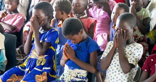 Children sitting together indoors with their heads bowed and hands clasped in prayer.