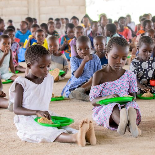 Children sitting on the ground eating a meal from green plates, with many others gathered behind them.