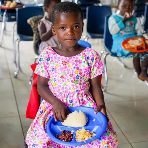 Young girl sitting on a chair holding a plate of food on her lap, with other children in the background.