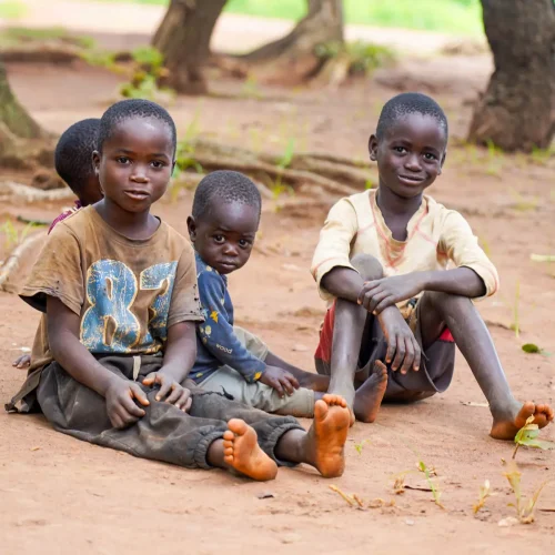 Four children sit barefoot on the ground in rural Zambia. Two boys in worn shirts look directly at the camera, while another child sits behind them. The group appears calm and thoughtful in their natural outdoor setting.